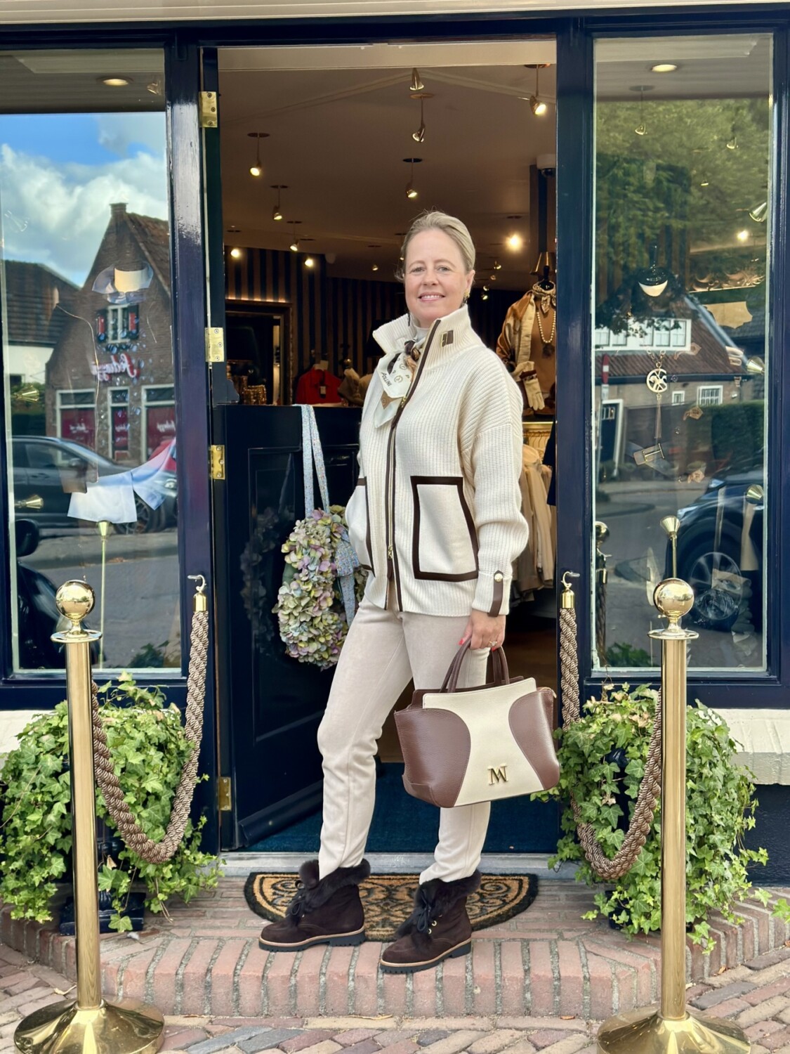 Elegant vrouw staat voor boutique Modici met stijlvolle beige en bruine kleding, accessoires en bagage, in een gezellige winkelstraat.
